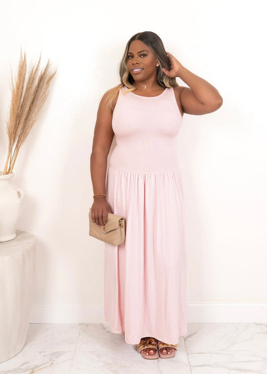 Plus-size model wearing a soft pink cotton candy maxi dress with a gathered waist, styled with a beige clutch and flat sandals — perfect dress from Opulent Gurlz.