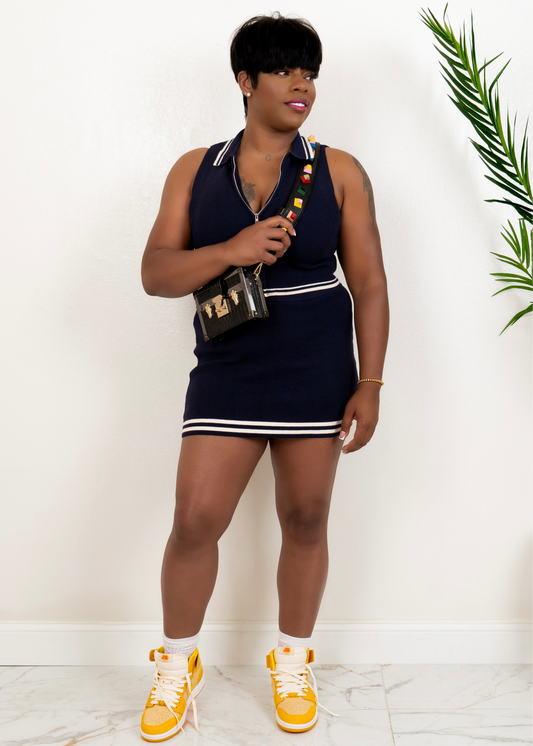 Front view of navy two-piece knit set with zip crop top and matching mini skirt, styled with sneakers and crossbody bag.
