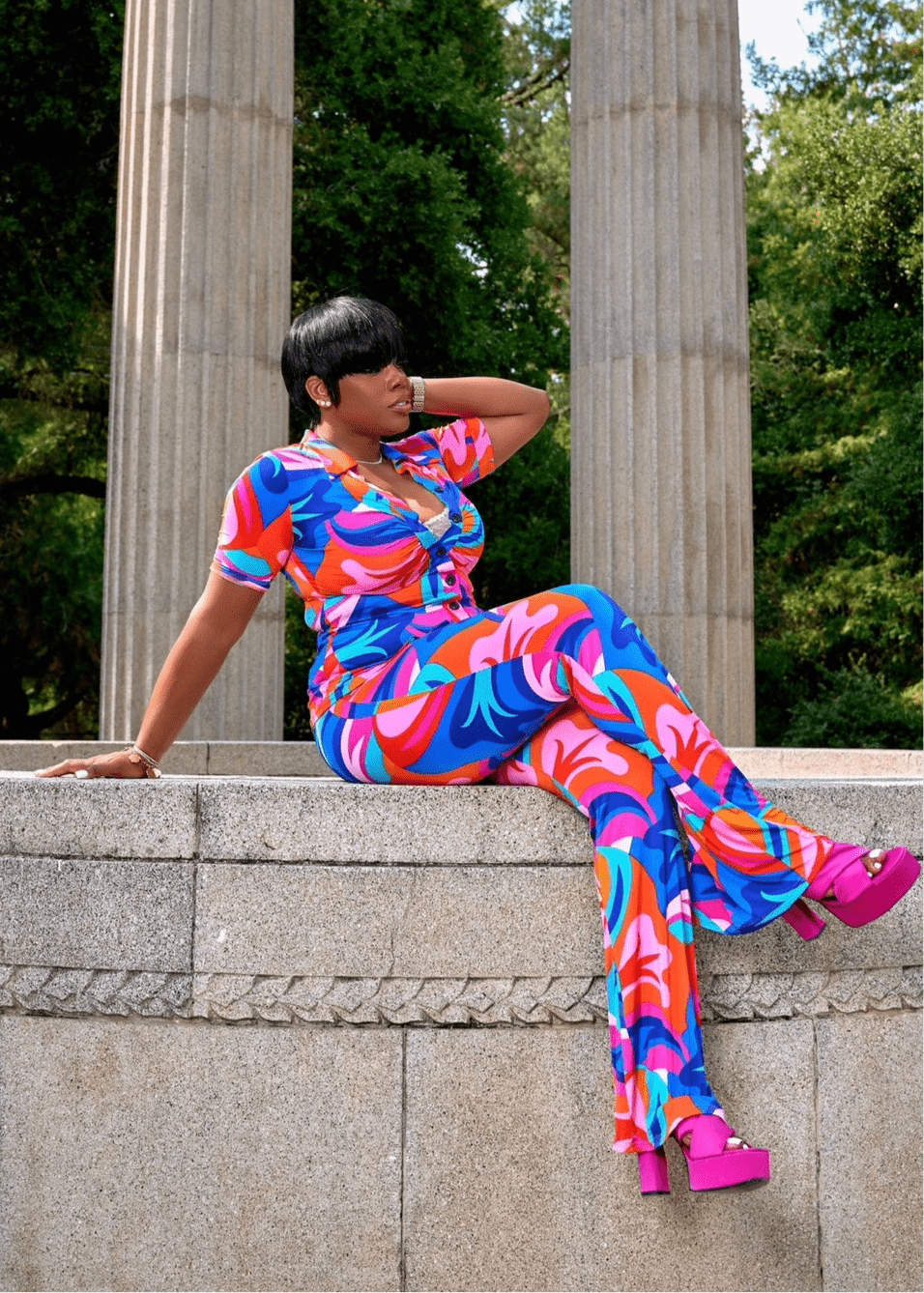 Model seated in vibrant abstract print outfit featuring wide-leg pants and a collared shirt, styled for summer glam near neoclassical columns.