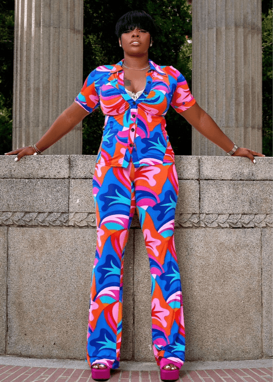 Model wearing bold multicolor vacation outfit with short-sleeve button top and wide-leg pants, styled with platforms in front of stone pillars.