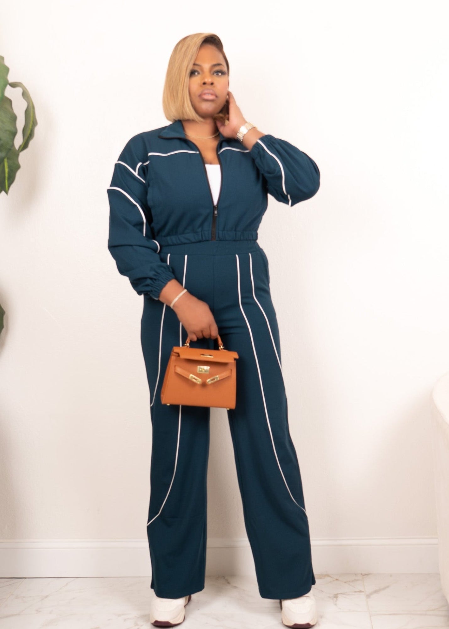 Model wearing hunter green and ivory contrast piping zip-up track set with wide-leg pants, full-body front view holding tan handbag