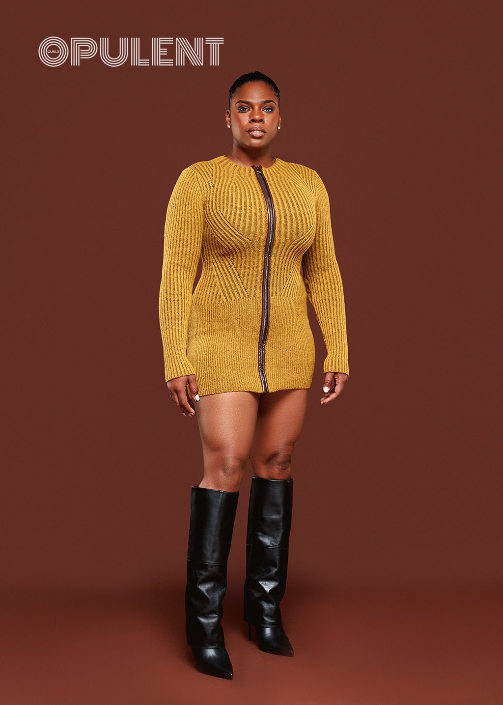 Model wearing green zip front sweater dress with long sleeves, leather detail, and black boots on red background