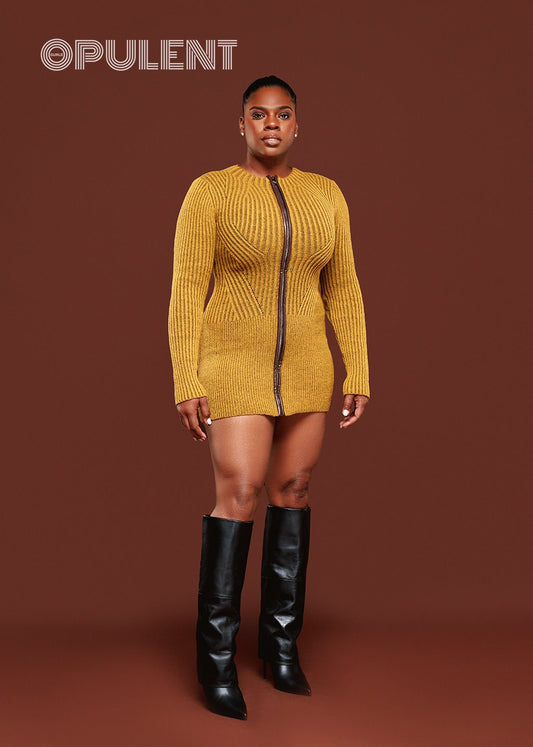 Model wearing green zip front sweater dress with long sleeves, leather detail, and black boots on red background
