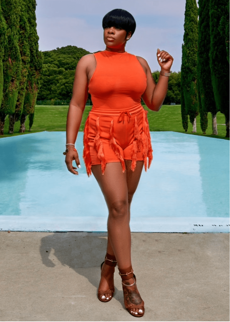Model wearing a vibrant orange fringe outfit featuring a sleeveless mock neck top and high-waisted tassel shorts, posing by a scenic poolside backdrop.
