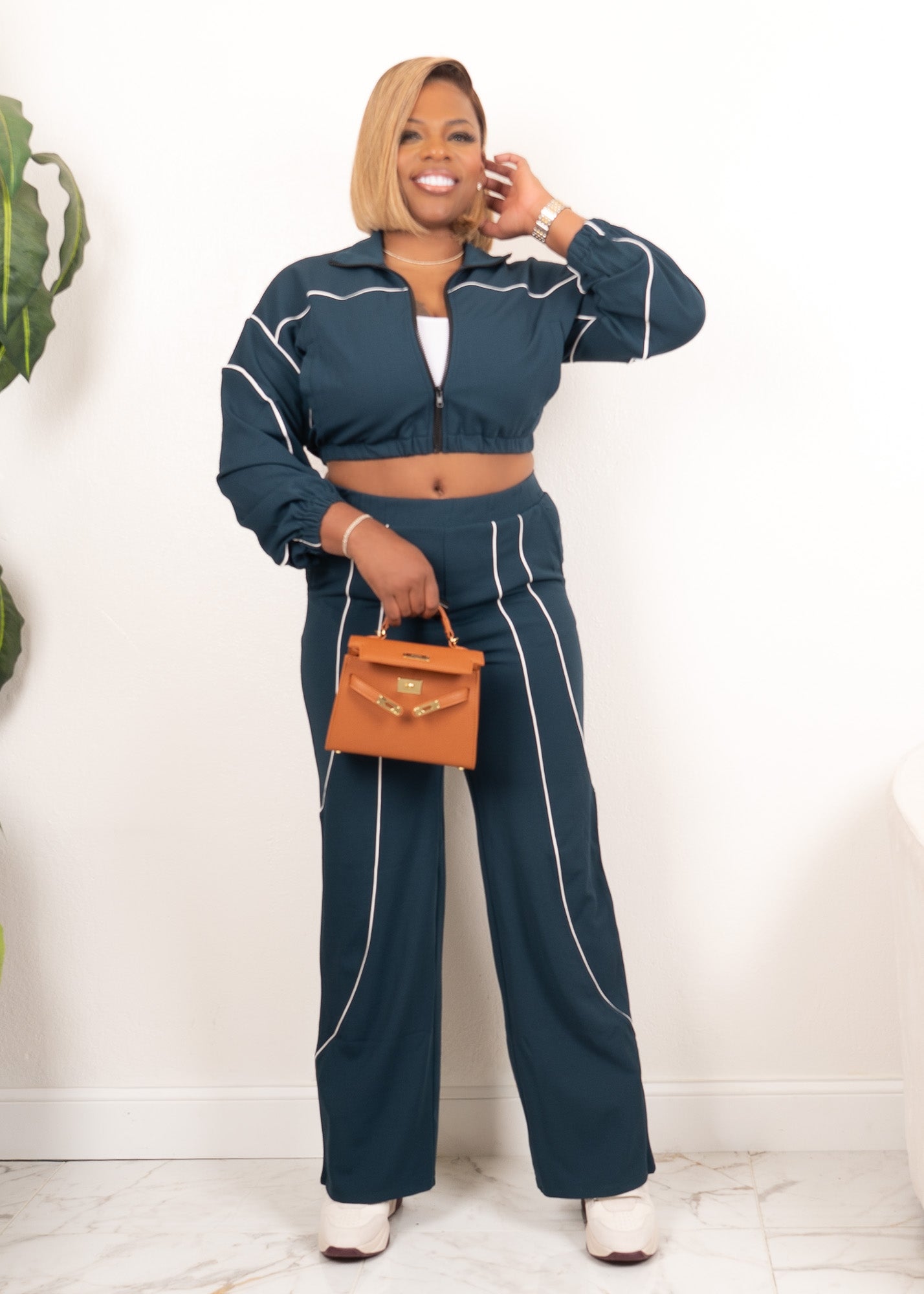 Model wearing hunter green and ivory contrast piping zip-up track set with wide-leg pants, full-body front view holding tan handbag